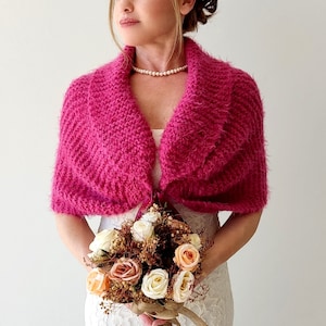 K&ouml;nnte beinhalten: Ein rosa gestrickter Schal mit einem breiten Kragen und einer Schleife vorne. Der Schal ist aus weichem, flauschigem Garn gefertigt und perfekt, um jedem Outfit einen Hauch von W&auml;rme und Stil zu verleihen.