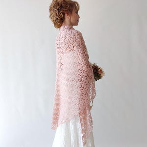Puede incluir: Un chal de crochet rosa claro con un dise&ntilde;o floral, usado sobre un vestido blanco. El chal est&aacute; drapeado sobre los hombros y tiene un tejido delicado y abierto. La imagen muestra a una mujer con el pelo recogido, sosteniendo un ramo de flores.