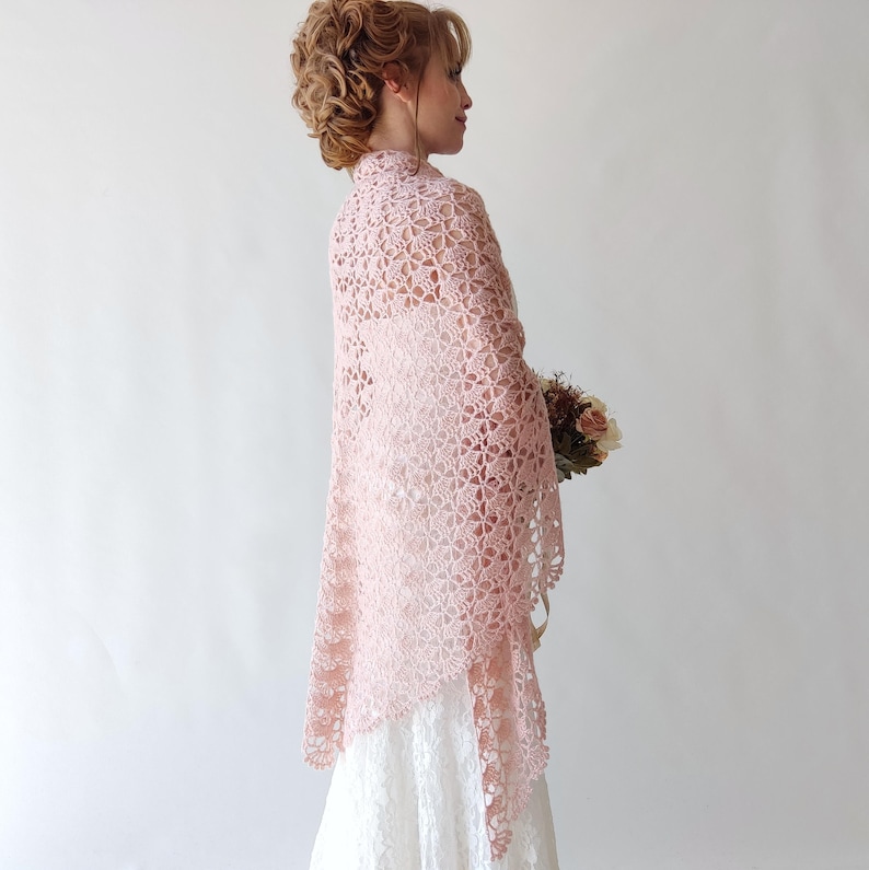 Puede incluir: Un chal de crochet rosa claro con un dise&ntilde;o floral, usado sobre un vestido blanco. El chal est&aacute; drapeado sobre los hombros y tiene un tejido delicado y abierto. La imagen muestra a una mujer con el pelo recogido, sosteniendo un ramo de flores.