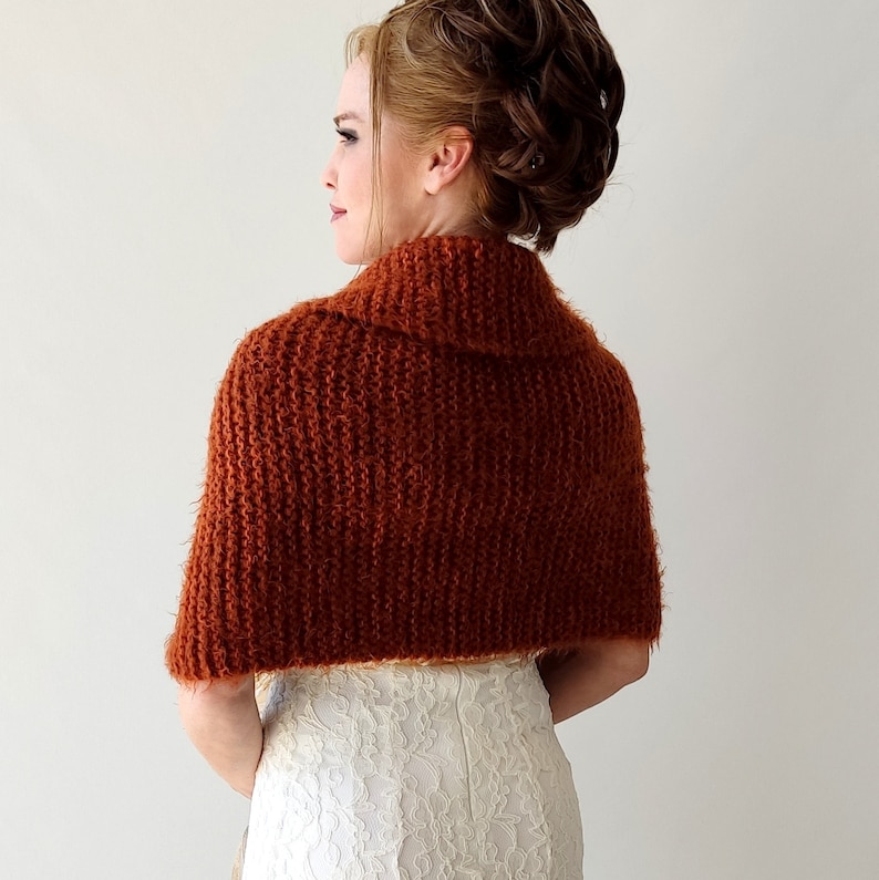 May include: A rust-colored, chunky knit shrug with a textured, fuzzy yarn finish. The garment features a wide, folded collar and covers the shoulders and upper back, worn over a white, floral-patterned lace dress with a visible back zipper.