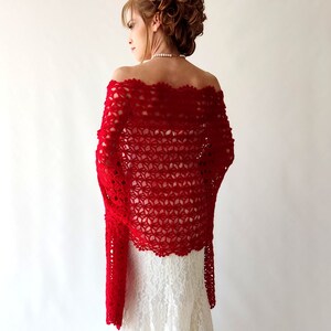 May include: A vibrant red, off-the-shoulder crochet shawl. The shawl features a delicate, openwork floral pattern and long, flowing sleeves. The garment is worn over a white dress, creating a striking contrast.
