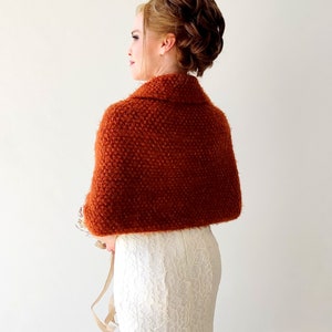 May include: A rust-colored, textured knit shawl with a folded collar draped over the shoulders, worn over a white, floor-length, lace-patterned dress. The shawl features a dense, nubby yarn construction and covers the upper back and arms.