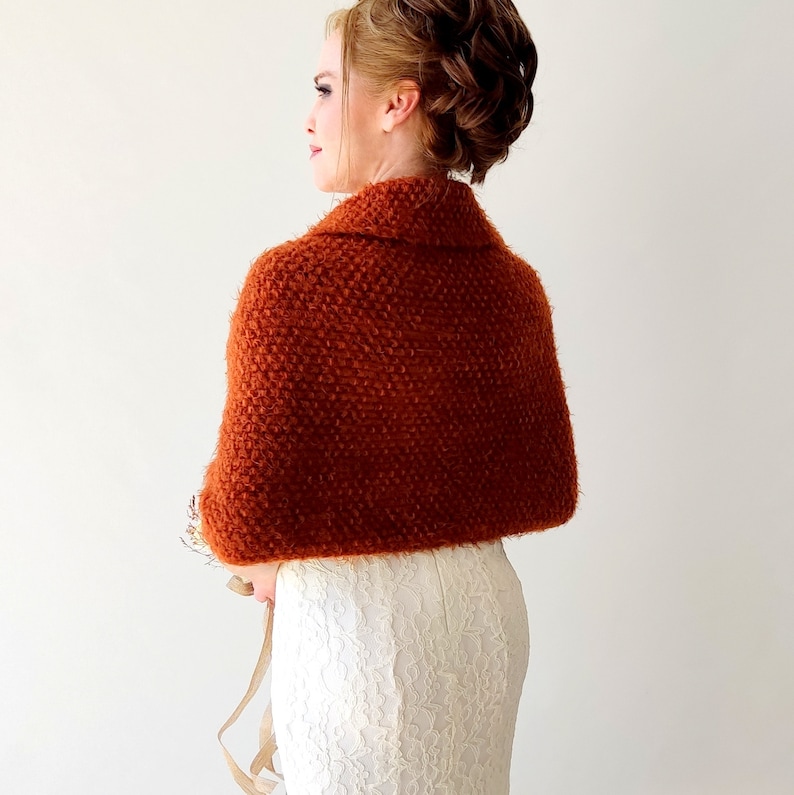 May include: A rust-colored, textured knit shawl with a folded collar draped over the shoulders, worn over a white, floor-length, lace-patterned dress. The shawl features a dense, nubby yarn construction and covers the upper back and arms.