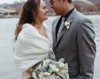 May include: A white knitted shawl draped over the shoulders of a person wearing a lace dress, standing next to a person in a grey suit with a dark shirt, a patterned tie, and a white floral boutonniere, holding a bouquet of white and purple flowers.
