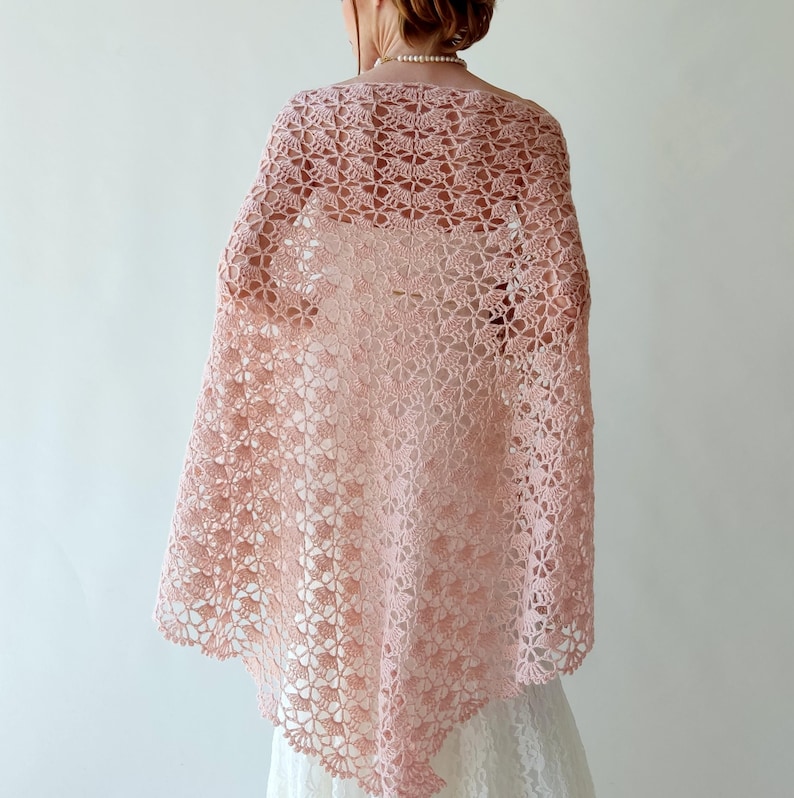 Puede incluir: Un chal de ganchillo rosa claro de tejido abierto. El chal est&aacute; drapeado sobre los hombros, mostrando un delicado estampado floral. El intrincado dise&ntilde;o y el color suave crean una est&eacute;tica elegante y aireada, adecuada para ocasiones especiales o para a&ntilde;adir un toque de encanto vintage a un atuendo.