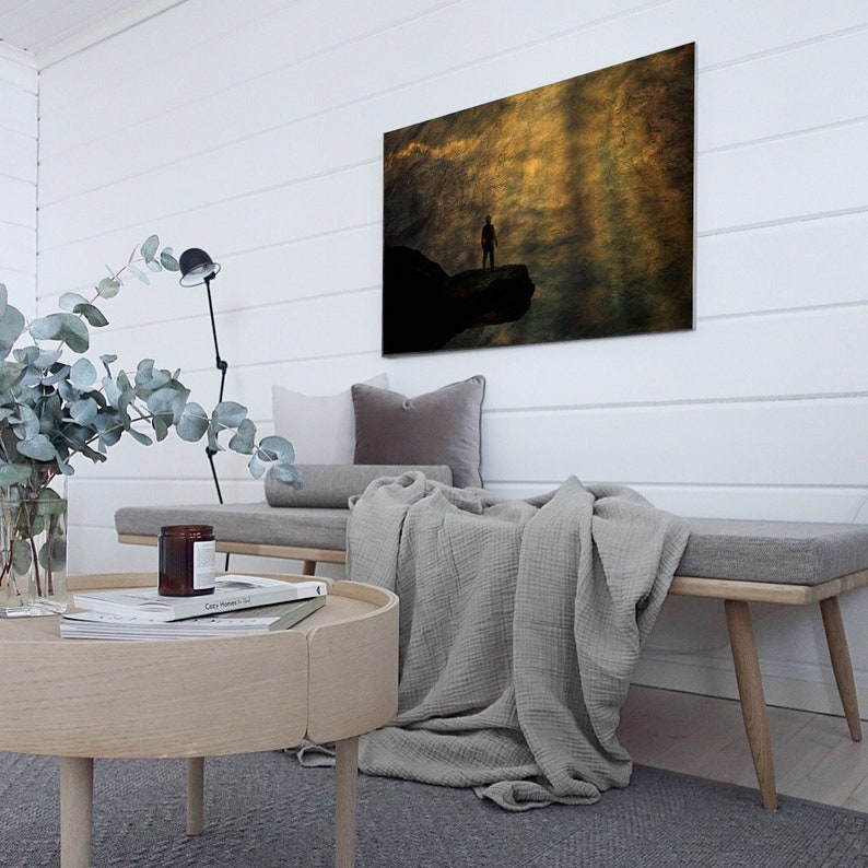 K&ouml;nnte beinhalten: Eine hellgraue Wolldecke liegt &uuml;ber einer Holzbank mit einem grauen Kissen. Ein Holz-Couchtisch mit einem Buch und einer Kerze steht vor der Bank. Ein gerahmtes Bild mit der Silhouette einer Person, die auf einem Felsen steht, vor einem goldenen Sonnenuntergang, h&auml;ngt an der Wand hinter der Bank.