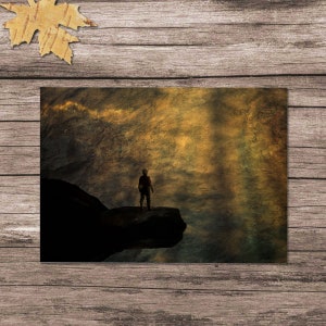 K&ouml;nnte beinhalten: Eine Silhouette einer Person, die auf einer Klippe steht und auf eine weite, goldene und strukturierte Landschaft blickt. Der Himmel ist gef&uuml;llt mit wirbelnden Wolken und einem sanften, &auml;therischen Schein.