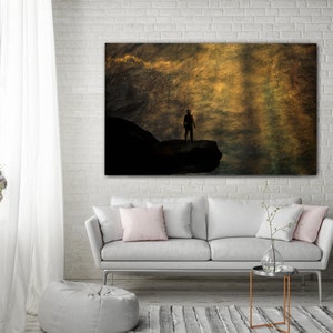 K&ouml;nnte beinhalten: Eine Silhouette einer Person, die auf einem felsigen Vorsprung steht, vor dem Hintergrund eines goldenen, strukturierten Himmels.