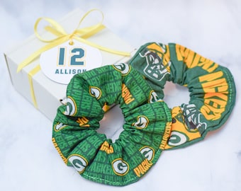 Puede incluir: Dos scrunchies de tela verde y dorada con un logotipo de equipo de fútbol y las palabras "Packers" y "Green Bay" impresas en ellos. Una caja de regalo blanca con una cinta amarilla y una etiqueta que dice "12 Allison" está en el fondo.