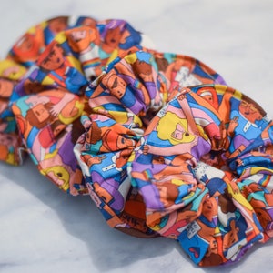Puede incluir: Un scrunchie para el pelo colorido con un patrón de personas que se toman de la mano. El scrunchie está hecho de tela y tiene un diseño de arcoíris.
