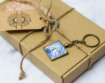 May include: A brown cardboard box with a brown twine bow and a small square keychain with a blue and white image inside. The keychain is attached to a silver chain and a bronze key ring. A brown tag with a sunflower and the word "Vincent" is attached to the box.