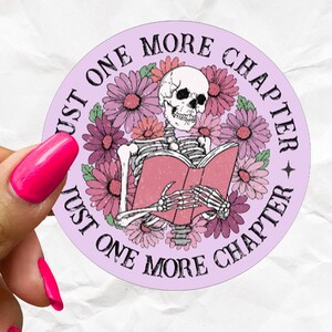 Op de afbeelding: Een ronde sticker met een lichtpaarse achtergrond met een skelet dat een roze boek leest, omringd door roze en paarse bloemen. De tekst "JUST ONE MORE CHAPTER" staat in zwarte hoofdletters rondom de cirkelvormige rand gedrukt.