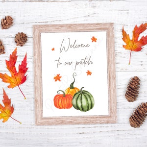 May include: A framed print with the text "Welcome to our patch" and a watercolour illustration of three pumpkins, two orange and one green, with autumn leaves scattered around the pumpkins. The print is on a white wooden background with autumn leaves and pine cones.
