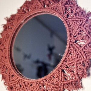 May include: A round mirror with a macrame frame in a dusty rose colour. The frame is made of thick cord and has a fringe around the edge.