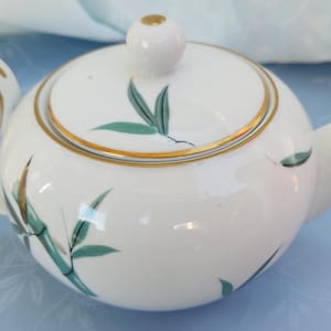 May include: White porcelain teapot with a gold trim and a green bamboo leaf design. The teapot has a lid with a knob on top.