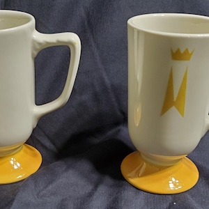 May include: Two white ceramic mugs with a yellow crown and banner design. The mugs have a yellow base and a curved handle. The mugs are suitable for coffee, tea, or other beverages.