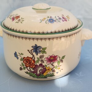 May include: A white ceramic casserole dish with a lid, featuring a floral pattern in shades of pink, blue, green, and yellow. The dish has a green trim around the top and two handles on the sides.