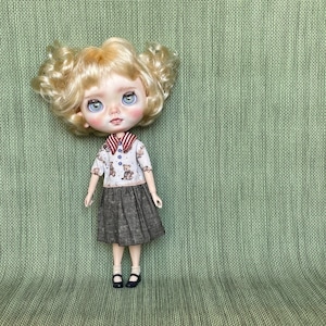 May include: A doll with curly blonde hair and large blue eyes wearing a white short-sleeved shirt with a brown teddy bear print, a red and white striped collar, and a dark brown skirt with a subtle grid pattern, paired with black shoes.
