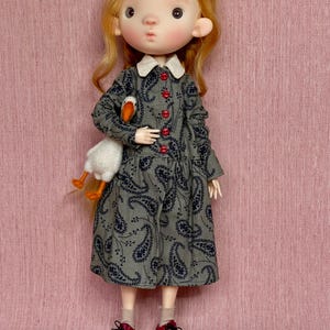May include: A doll with auburn hair wearing a gray paisley dress with red buttons and shoes. The doll is holding a white stuffed goose with orange feet and beak. The dress has a white collar.