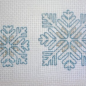 Puede incluir: Dos copos de nieve bordados sobre un fondo de tela blanca. El copo de nieve más grande es azul y dorado, mientras que el copo de nieve más pequeño es azul.