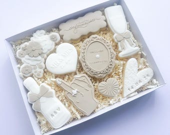 May include: A selection of wedding-themed biscuits in a white box. The biscuits are decorated with white and beige icing and include shapes such as a champagne bottle, a champagne glass, and a heart. The box is filled with shredded paper.