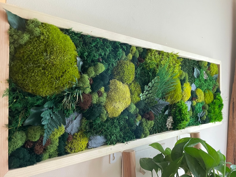 May include: A rectangular wall art piece featuring a vibrant arrangement of preserved moss and various plants in shades of green, brown, and yellow. The artwork is encased in a light-colored wooden frame, creating a natural and textured aesthetic. The moss art adds a touch of nature to any interior space.