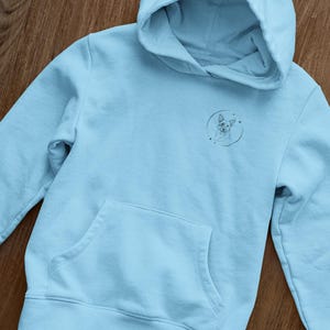 K&ouml;nnte beinhalten: Ein hellblauer Hoodie mit einem kleinen, schwarz-wei&szlig;en Grafikdruck eines Hundekopfes in einem Kreis auf der Vorderseite. Der Hoodie hat eine K&auml;ngurutasche und eine Kapuze mit Kordelzug.