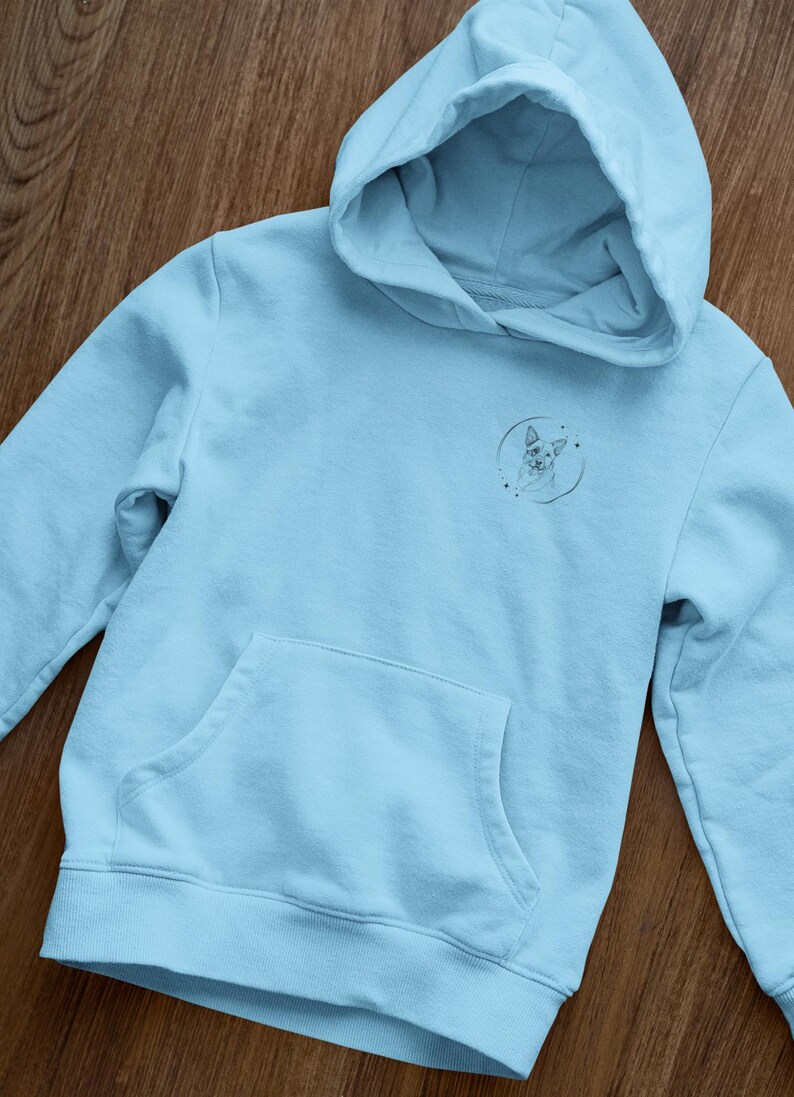 K&ouml;nnte beinhalten: Ein hellblauer Hoodie mit einem kleinen, schwarz-wei&szlig;en Grafikdruck eines Hundekopfes in einem Kreis auf der Vorderseite. Der Hoodie hat eine K&auml;ngurutasche und eine Kapuze mit Kordelzug.