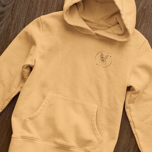 K&ouml;nnte beinhalten: Ein hellgelber Hoodie mit einer kleinen schwarz-wei&szlig;en Grafik eines Hundekopfes in einem Kreis auf der linken Brust.