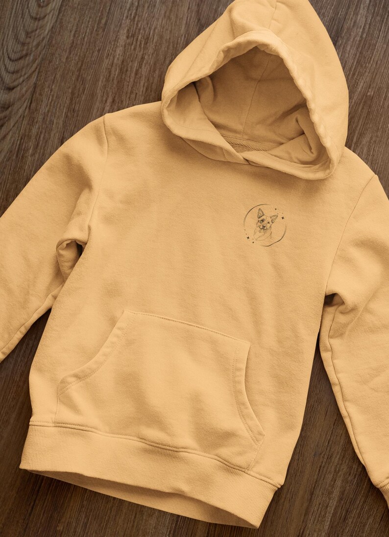K&ouml;nnte beinhalten: Ein hellgelber Hoodie mit einer kleinen schwarz-wei&szlig;en Grafik eines Hundekopfes in einem Kreis auf der linken Brust.