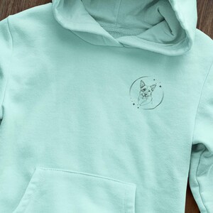 K&ouml;nnte beinhalten: Ein hellblauer Hoodie mit einer kleinen schwarz-wei&szlig;en Illustration eines Hundekopfes in einem Kreis auf der Vorderseite.