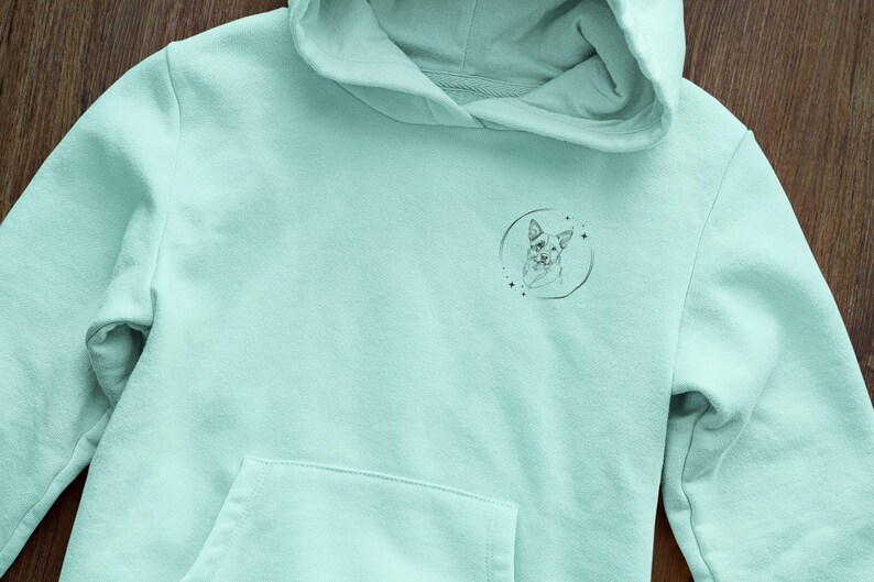 K&ouml;nnte beinhalten: Ein hellblauer Hoodie mit einer kleinen schwarz-wei&szlig;en Illustration eines Hundekopfes in einem Kreis auf der Vorderseite.