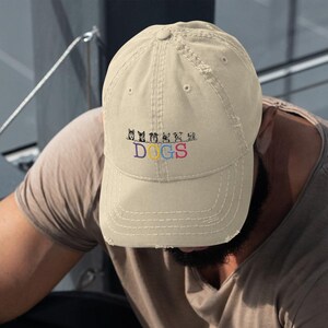 Peut inclure: Une casquette de baseball beige, usée, avec un motif brodé de cinq chiens de dessin animé avec le texte "EMOTIONAL DOGS" en lettres colorées.