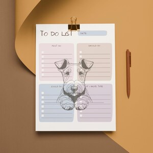 May include: A white To Do List notepad with a dog illustration, held by a gold clip. The notepad has sections for Must Do, Should Do, Could Do, and If I Have Time. A brown pen rests beside the notepad.