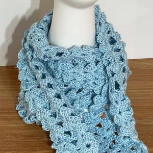 May include: A light blue crocheted shawl displayed on a mannequin head. The shawl is made of a soft, open-weave yarn, with a floral pattern. The color is a soft, pastel blue. The shawl is draped around the mannequin's neck and shoulders.
