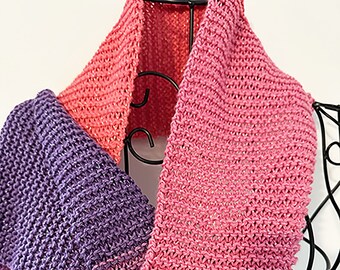 May include: A knitted cowl scarf in shades of pink and purple. The scarf is displayed on a black metal stand. The colors transition from a bright pink at the top, fading into a darker purple at the bottom. The knit pattern is visible.