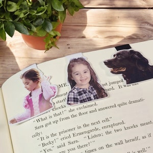 May include: Three decorative bookmarks are inserted into a book. The bookmarks feature images of a young girl in a pink dress, a smiling girl in a plaid shirt, and a black dog. The book is open on a wooden surface.