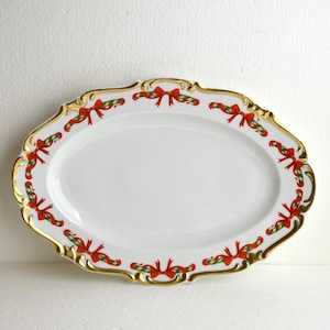 以下が含まれることがあります： 金色の縁と赤と緑のリボンのデザインが施された白い楕円形のサービングプラッター。お祝いに最適です。前菜やデザートのサービングに最適なクラシックなアイテムです。