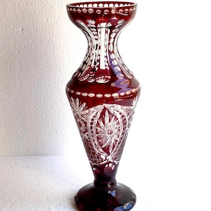 May include: A tall, ruby red crystal vase with intricate etched floral and geometric patterns. The vase has a flared top, a slender neck, and a wide, round base. The design includes a series of small, circular cutouts around the rim.