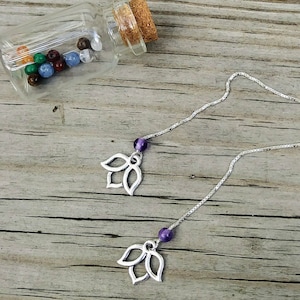 Puede incluir: Pendientes de cadena plateada con un dije de flor de loto y una cuenta morada. Una pequeña botella de vidrio con tapón de corcho contiene cuentas de colores. Los pendientes se exhiben sobre una superficie de madera.
