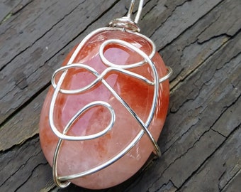 May include: A handcrafted pendant featuring a polished, oval-shaped, reddish-orange gemstone wrapped in a silver-colored wire. The wire is intricately woven around the stone, creating a decorative design with a loop at the top for a chain.