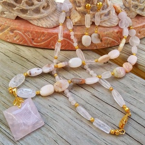 May include: A necklace and earring set featuring a variety of light pink and white gemstones. The necklace has a square pendant and gold-colored accents. The earrings have a matching design. The jewelry is displayed on a wooden surface.