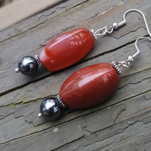 May include: A pair of earrings featuring elongated, reddish-brown beads paired with a dark gray, spherical bead. The earrings have silver-colored hooks and decorative metal accents.