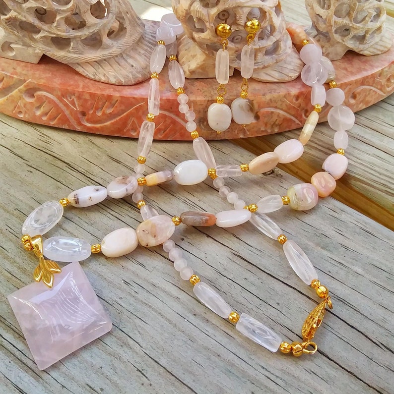 May include: A necklace and earring set featuring a variety of light pink and white gemstones. The necklace has a square pendant and gold-colored accents. The earrings have a matching design. The jewelry is displayed on a wooden surface.