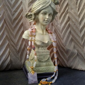May include: A bust of a woman with a rose in her hair is adorned with a pink and white beaded necklace. A square, lavender pendant with a gold leaf design and a matching necklace with a gold clasp are also present. The background is a textured, light brown fabric.
