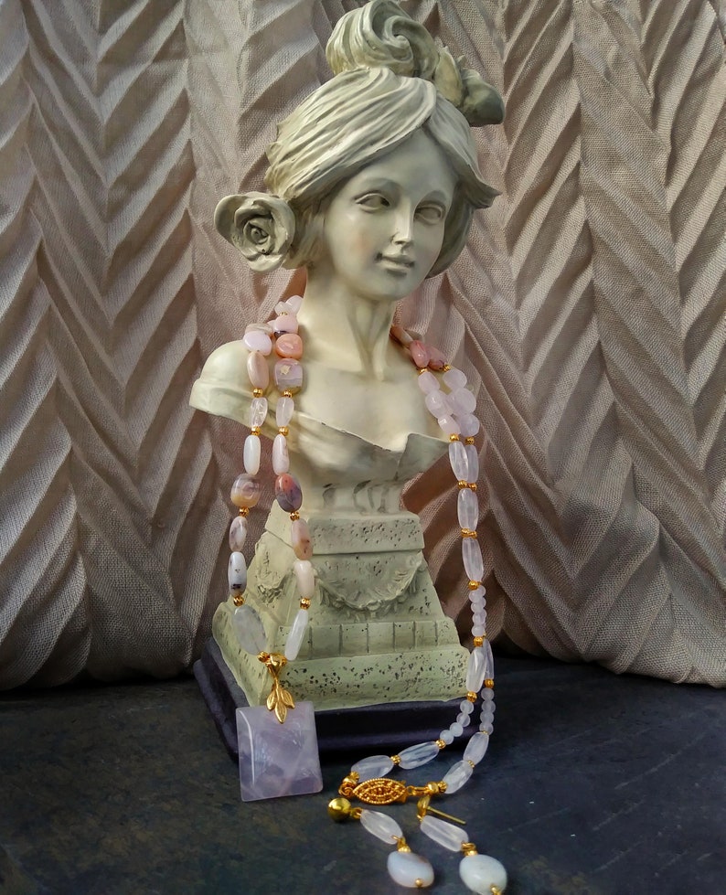 May include: A bust of a woman with a rose in her hair is adorned with a pink and white beaded necklace. A square, lavender pendant with a gold leaf design and a matching necklace with a gold clasp are also present. The background is a textured, light brown fabric.