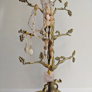 May include: A bronze-colored jewelry tree displays necklaces and earrings. The necklaces feature light pink and clear gemstone beads. A square rose quartz pendant hangs from the tree. The earrings are oval-shaped and dangle from the branches. The jewelry is displayed against a white background.
