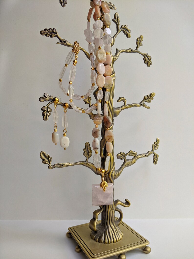 May include: A bronze-colored jewelry tree displays necklaces and earrings. The necklaces feature light pink and clear gemstone beads. A square rose quartz pendant hangs from the tree. The earrings are oval-shaped and dangle from the branches. The jewelry is displayed against a white background.