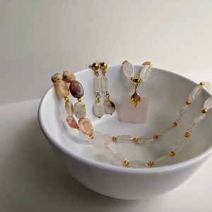 May include: A collection of gemstone jewelry, including a necklace, earrings, and a pendant necklace, displayed in a white bowl. The necklace features a mix of pink, beige, and brown stones. The pendant is a square rose quartz with a gold-colored leaf design.