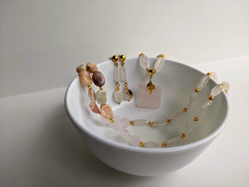 May include: A collection of gemstone jewelry, including a necklace, earrings, and a pendant necklace, displayed in a white bowl. The necklace features a mix of pink, beige, and brown stones. The pendant is a square rose quartz with a gold-colored leaf design.